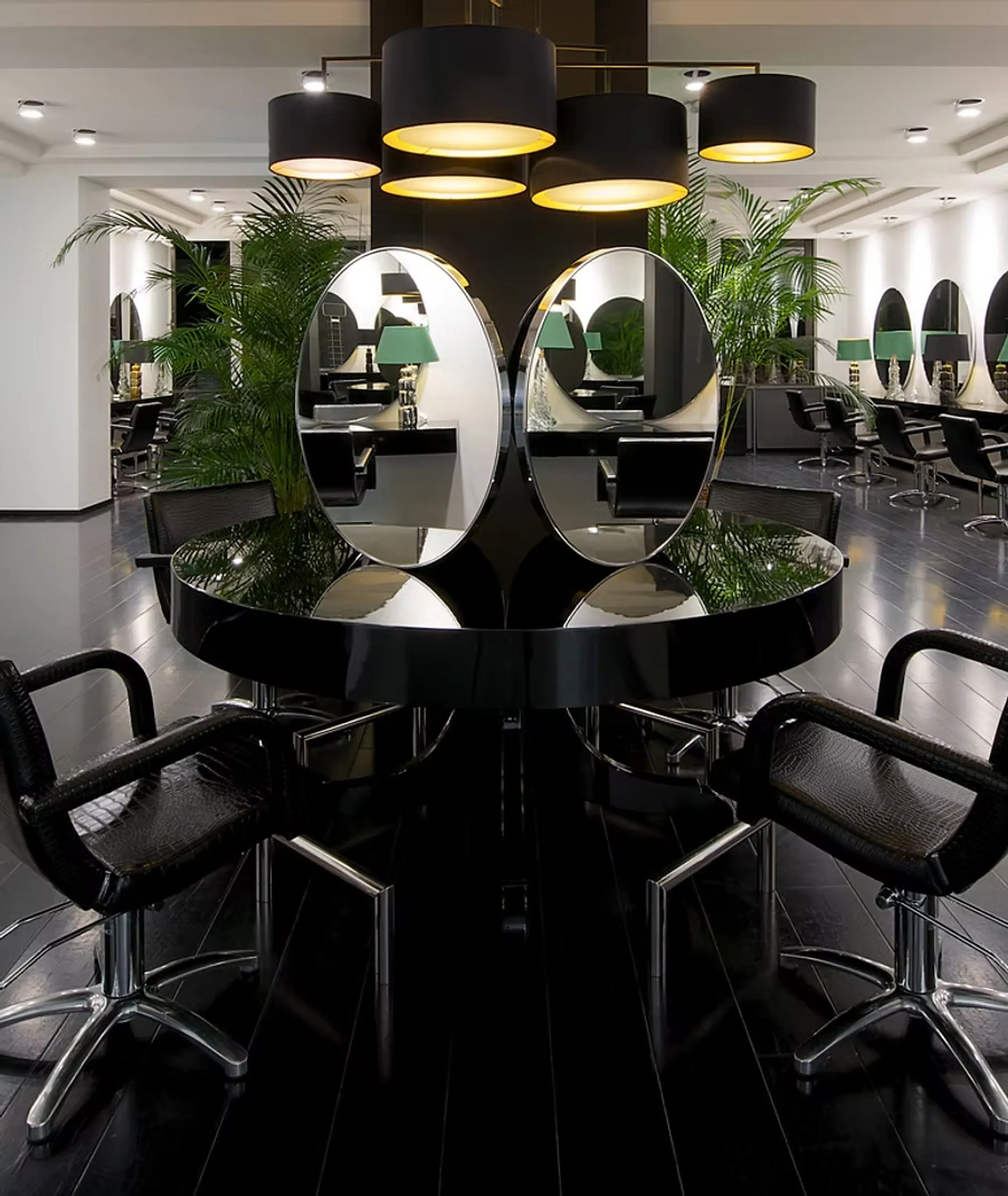Salonfläche Königsallee: runde schwarze Mittelinsel mit ovalen Doppelspiegeln, schwarze Stylingstühle, Palmen und schwarze Pendelleuchten mit goldenem Schirm