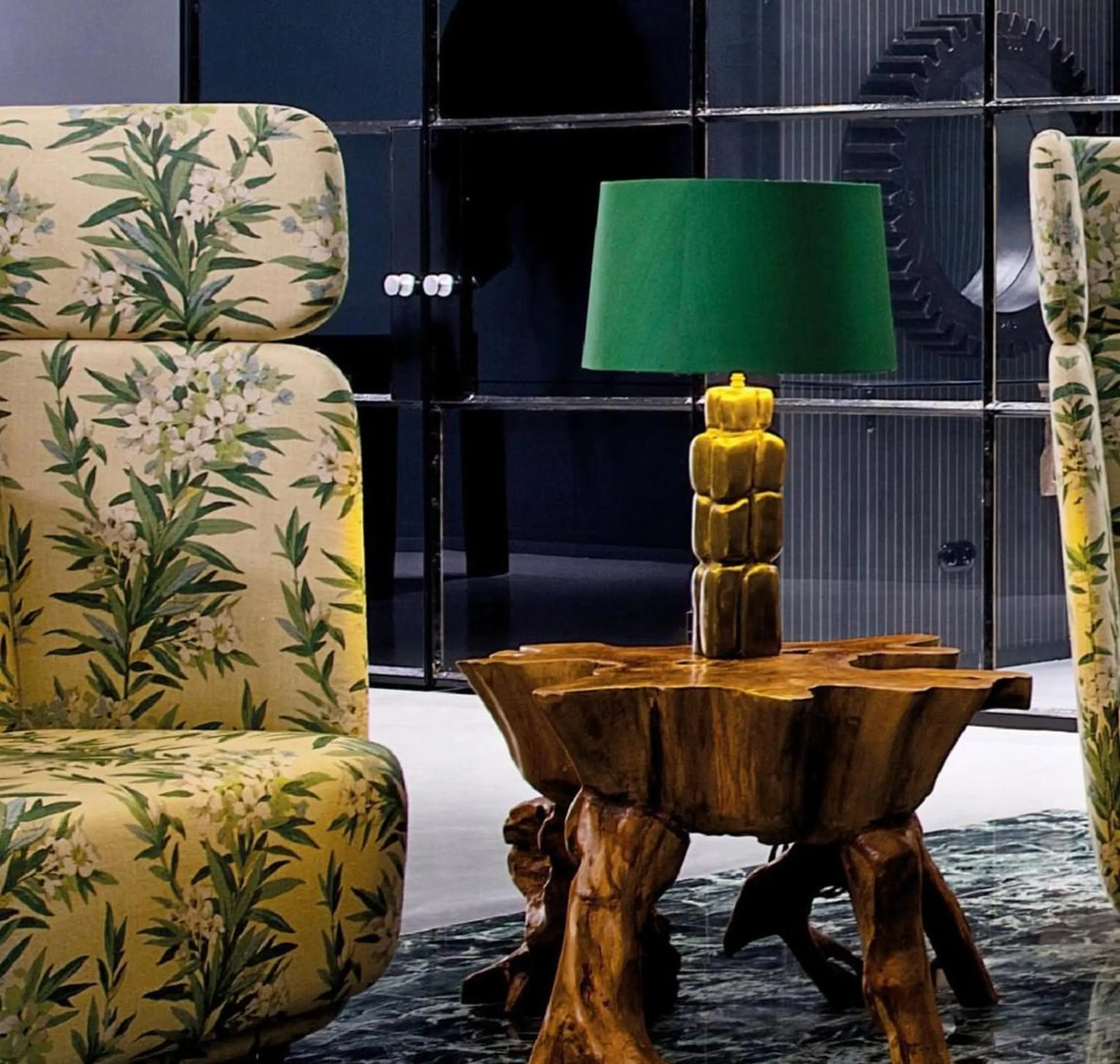 Lounge-Detail: geblümte Ohrensessel und Wurzelholztisch mit goldener Lampe mit grünem Schirm vor rauchverspiegelter Wand