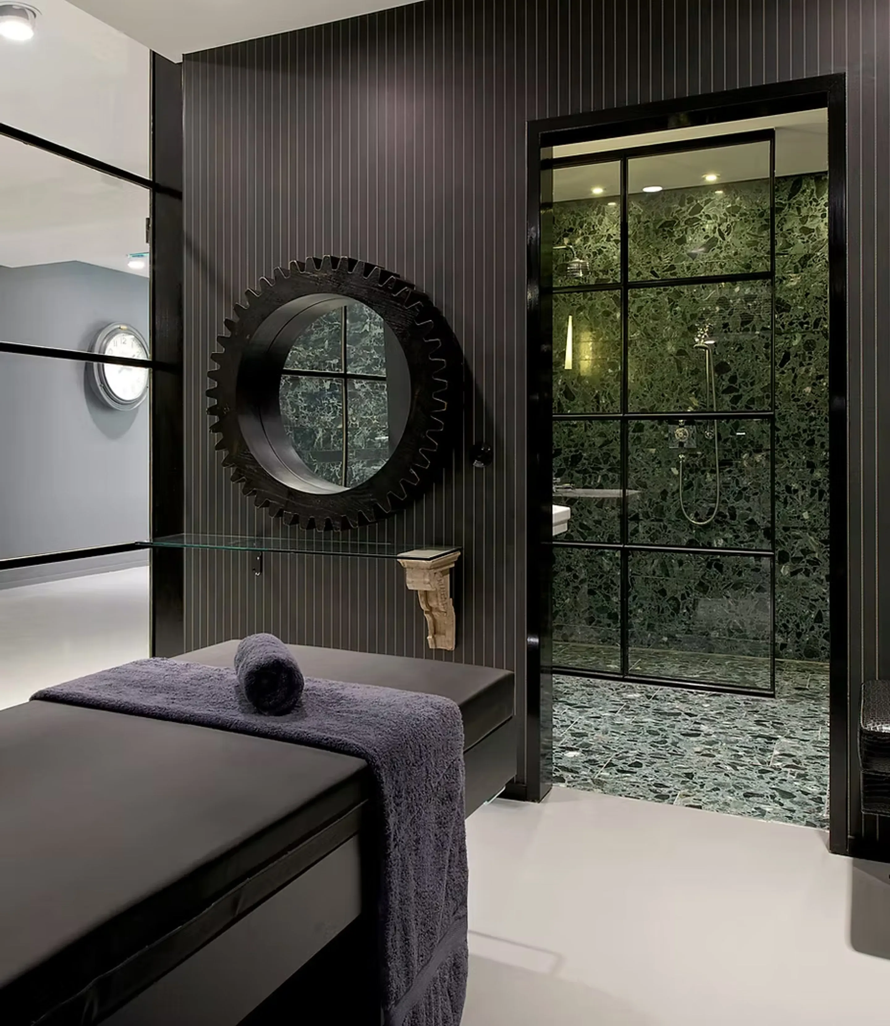 Spa-Bereich: schwarze Massageliege mit dunklen Handtüchern, zahnradförmiger Spiegel an Nadelstreifenwand, Blick in grün marmorierten Duschraum