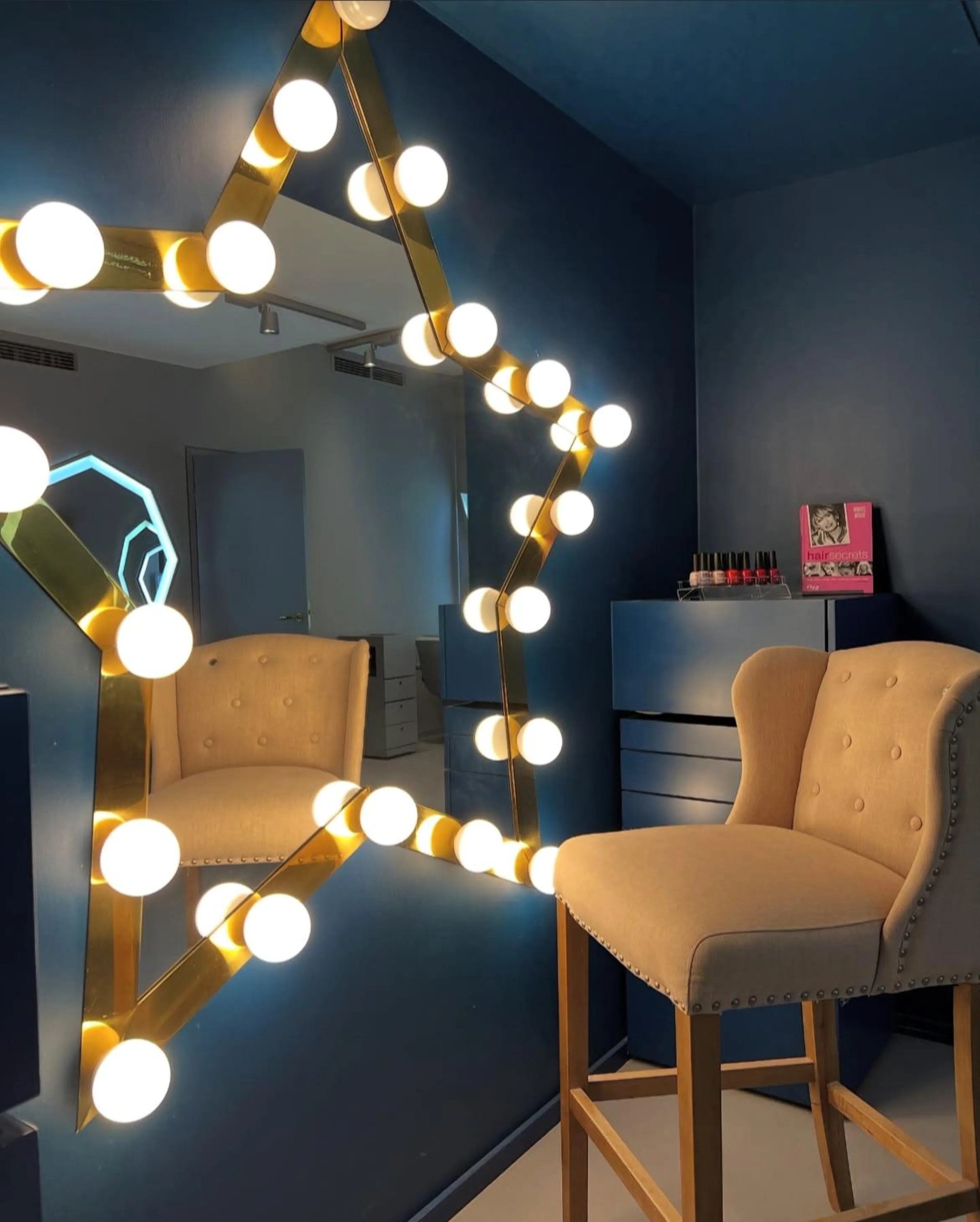 Beauty-Ecke im Salon Waitzstraße: sternförmiger Spiegel mit Glühbirnenkranz auf dunkelblauer Wand, heller Polsterstuhl und Nagellack-Display