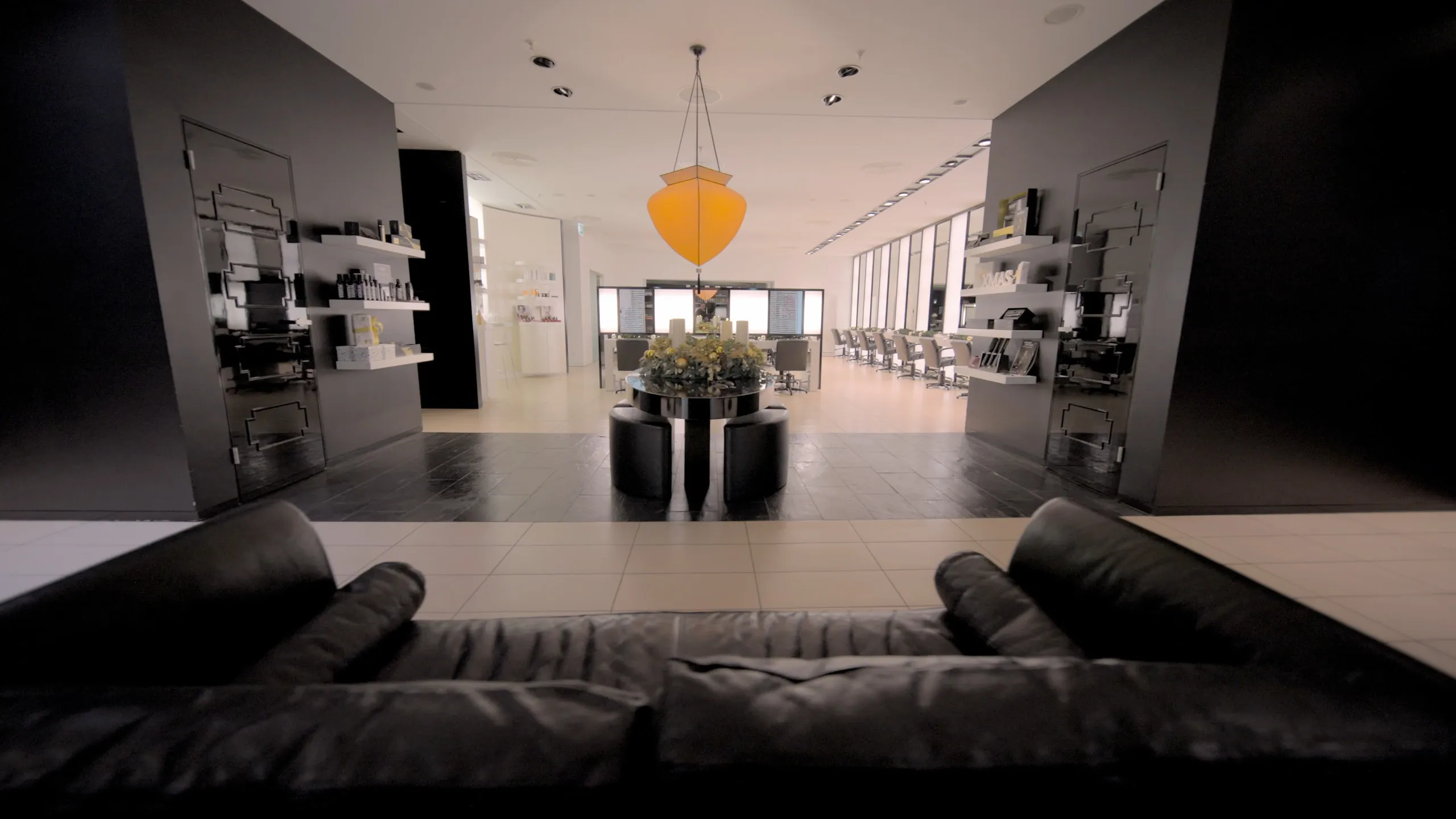 Blick über ein schwarzes Ledersofa in den Salon Alstertal mit orangefarbener Pendelleuchte, rundem Blumentisch und langen Stylingzeilen